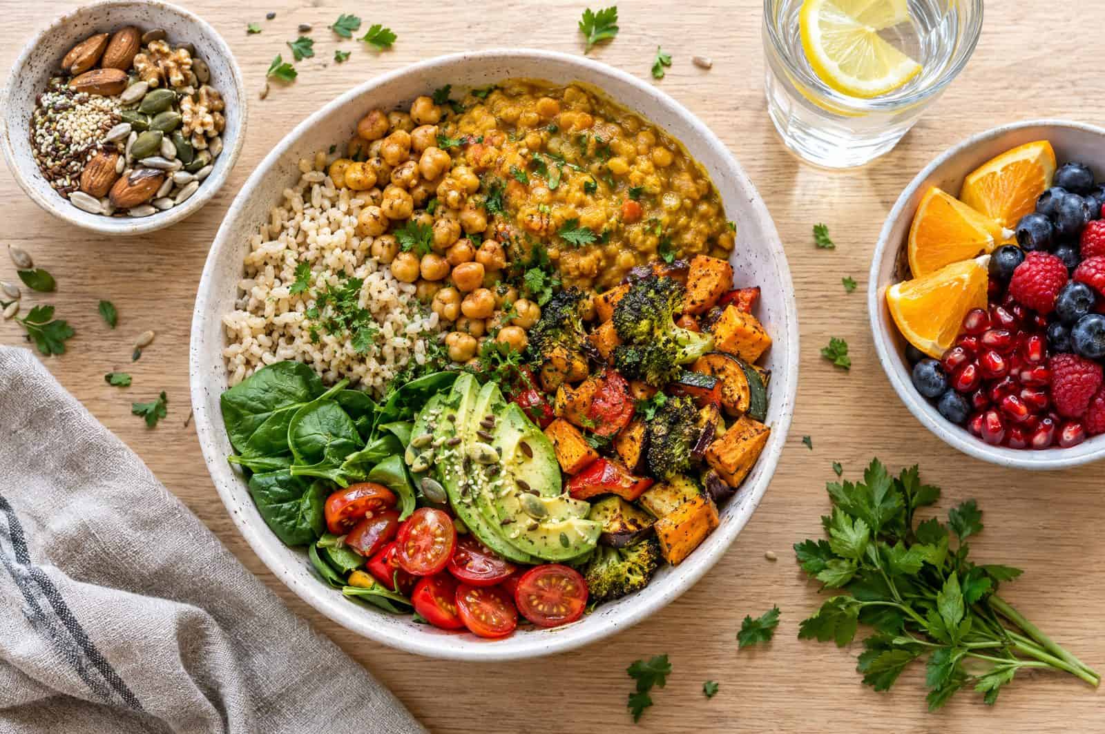 Colourful plant-based lunch bowl with brown rice, chickpeas, dal, roasted vegetables and avocado, with fruit and nuts on the side